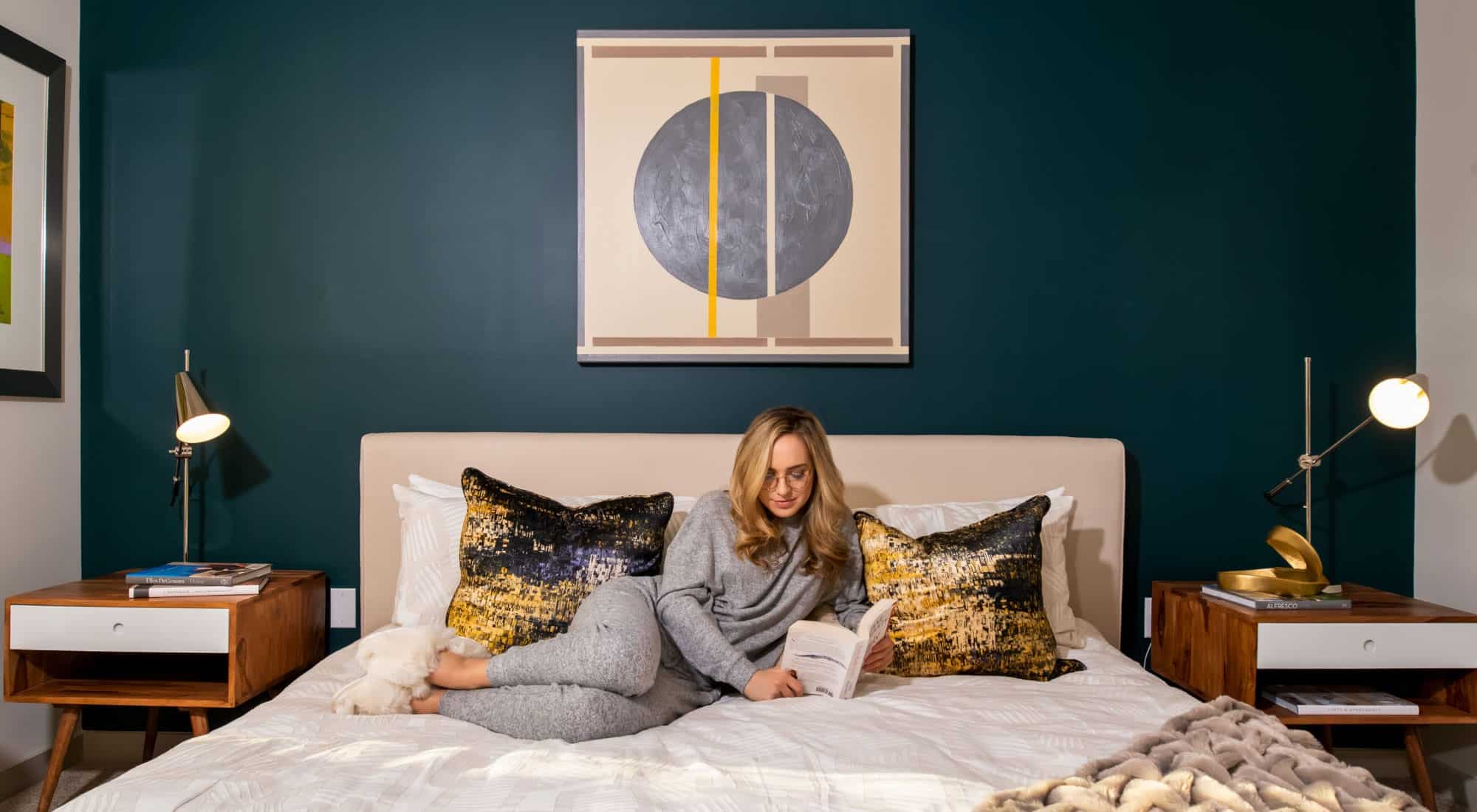 A blonde woman reads a book on a bed with a teal accent wall